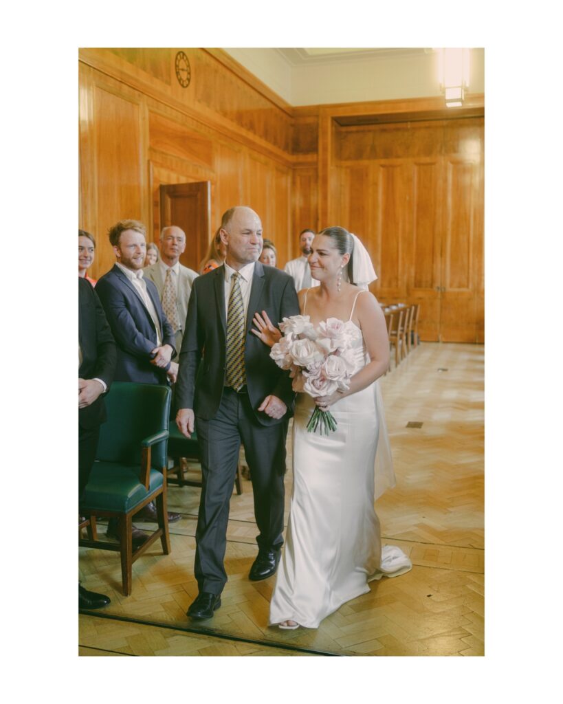 Bride emotionally walking down the aisle in arms with her Father who looks at her with love and pride. At Hackney Hall, London. Photo by London Wedding Photographer, Pia Bacino