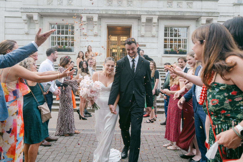 Couple walking out of Hackney Hall after getting married and all their guests tossing flower petals as they walk past. Photo by London Wedding Photographer, Pia Bacino